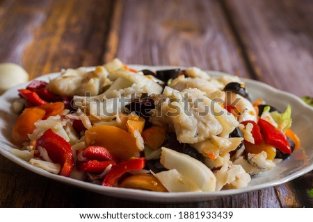 cod boiled with winter cherry tomatoes, black olives, celery, onion, pickled peppers on wooden background. Selective focus.
