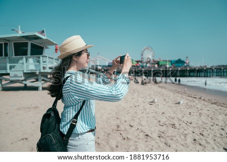 Travel lifestyle concept. side view beautiful asian chinese female tourist in straw hat and sunglasses taking photo on mobile phone while stand on sandy beach. girl traveler photographing cellphone