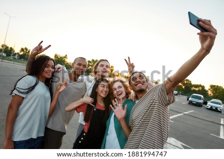 Group photo. Six young well-dressed friends having a good time together hugging each other smiling and making a group selfie outside on a parking site with cars on a background during a good weather