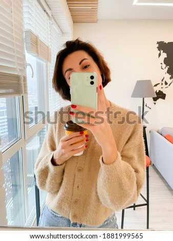 Young pretty short hair woman in cozy apartment at home in casual warm clothes hold yellow paper cup of coffee take photo selfie in mirror on mobile phone for stories and posts in social media
