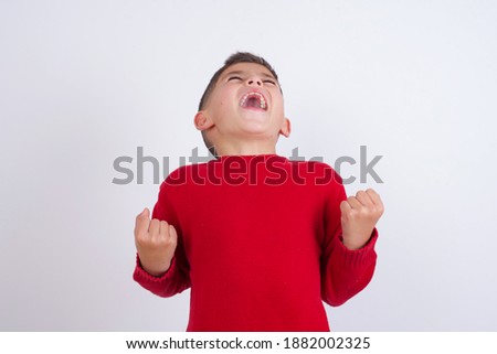 Cute Caucasian kid boy wearing knitted sweater against white wall looks with excitement up, keeps hands raised, notices something unexpected.
