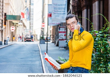Young Hispanic American Man with hair bun, wearing glasses, yellow long sleeve T shirt, sitting by green plants on old street in Manhattan, New York. reading red book, talking on cell phone.