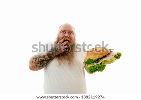 Isolated portrait on a white background of a fat man covering hid mouth with one hand holding and a huge burger on the other hand