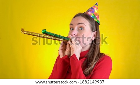 Young woman in birthday hat that is blowing in pipe, celebrating, over yellow background.