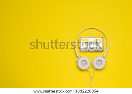 A gray cassette with magnetic tape and white headphones on a yellow background.
