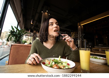 Young woman enjoys tasty meal. Attractive woman with brown hair slowly eating healthy caesar salad and looking away. Glass of orange juice. Restaurant or cafe on background. Cozy atmosphere. Royalty-Free Stock Photo #1882299244