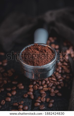 Tamped puck of coffee grounds within basket of portafilter and coffee beans spilled around in a dark and moody scene of natural light. Royalty-Free Stock Photo #1882424830