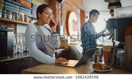 Latin American Coffee Shop Employee Accepts a Pre-Order on a Mobile Phone Call and Writes it Down on Laptop Computer in a Cozy Cafe. Restaurant Manager Browsing Internet and Talking on Smartphone. Royalty-Free Stock Photo #1882428598