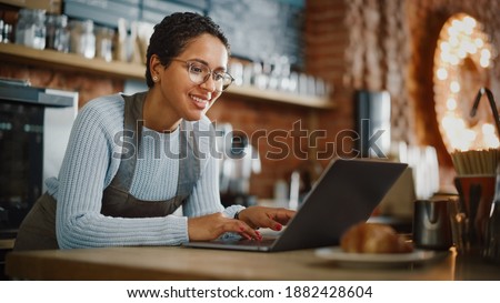 Young and Beautiful Latina Coffee Shop Owner is Working on Laptop Computer and Checking Inventory in a Cozy Cafe. Successful Restaurant Manager Browsing Internet and Accepting Online Take Away Orders. Royalty-Free Stock Photo #1882428604