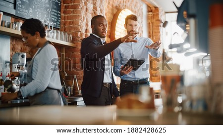 Multicultural Coffee Shop Owners Meeting Behind the Counter and Working on Tablet Computer and Checking Inventory in a Cozy Loft-Style Cafe. Successful Restaurant Managers and Barista at Work. Royalty-Free Stock Photo #1882428625