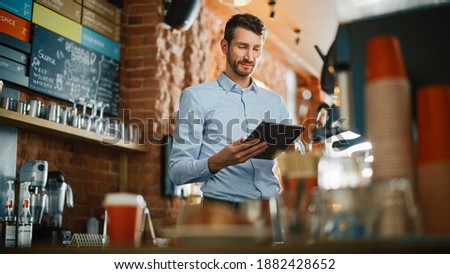 Handsome Caucasian Coffee Shop Owner is Working on Tablet Computer and Checking Inventory in a Cozy Loft-Style Cafe. Successful Restaurant Manager Standing Happy Behind Counter. Royalty-Free Stock Photo #1882428652