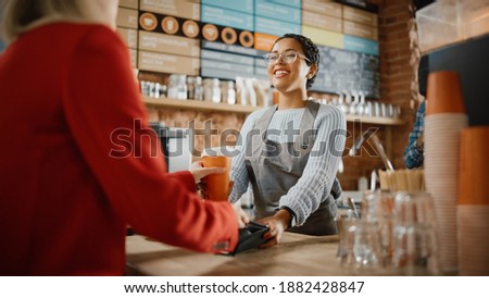 Joyful Multiethnic Diverse Woman Gives a Payment Terminal to Customer Using NFC Technology on Smartphone. Customer Uses Mobile to Pay for Take Away Latte and Croissant to a Barista in Coffee Shop. Royalty-Free Stock Photo #1882428847