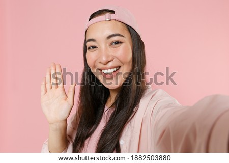 Close up of smiling young asian woman 20s in casual clothes cap isolated on pink background. People lifestyle concept. Mock up copy space. Doing selfie shot on mobile phone waving greeting with hand