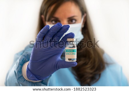 Caucasian female woman doctor or nurse in uniform and gloves wearing face mask protective in lab hold vaccine bottle and on bottle has "COVID-19 VACCINE" text label. Focus on text label.