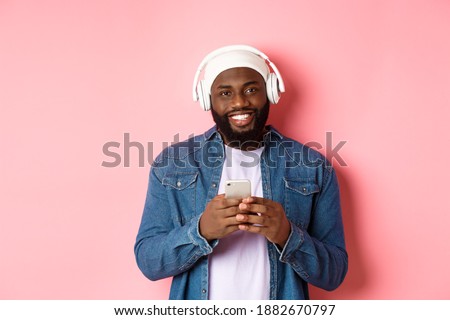 Image of handsome Black guy in headphones, listening music and using mobile phone, smiling at camera, pink background
