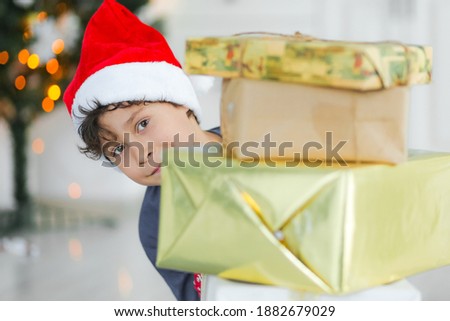 curly haired boy in santa claus hat holding gifts at home with christmas decor