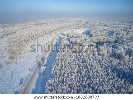 Aerial photo of winter road surrounded by birch forest. Drone shot of trees covered with hoarfrost and snow. Natural winter background.