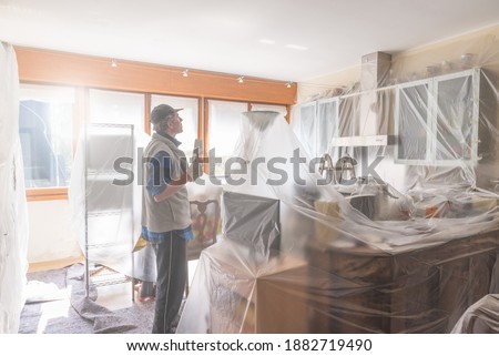 House painter painting the interior of a furnished house. Home renovation concept. Man is painting a kitchen protected with cover film Royalty-Free Stock Photo #1882719490