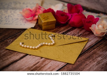 closed envelope with a pearl necklace on top and a gift box in the background with roses and old letters