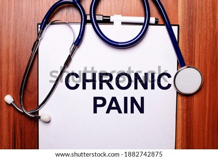 The word CHRONIC PAIN is written on white paper on a wooden background near a stethoscope and black-framed glasses. Medical concept