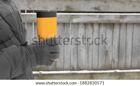 A man in gray clothes and gray gloves holds a thermos of bright yellow color. Wooden background with copy space.
