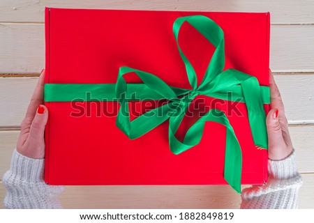Big Christmas Gift in Woman Hands. Large Red Christmas Gift Box with Festive Green Ribbon, girls hands in pics, top view on wooden background