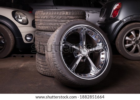 Automobile composition made up stack of tires and wheel with shiny metal disc in the foreground against the background of cars in a car service before seasonal replacement or after breaking through. Royalty-Free Stock Photo #1882851664