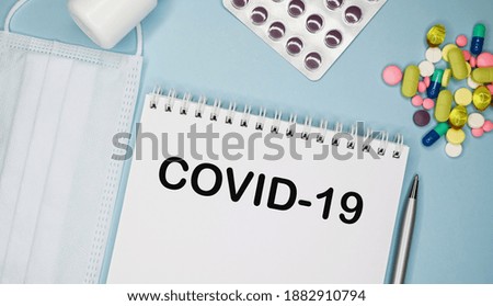 COVID 19 is written in a notebook on a blue background next to pills, mask and pen.