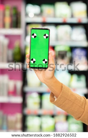 Hand of female shopaholic holding smartphone with green e-coupon on screen in front of camera against display with beauty products
