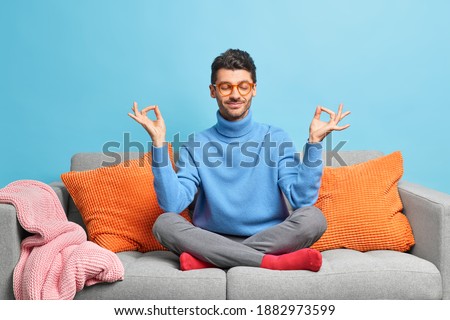 Calm relaxed man sits in lotus pose meditates at home during quarantine poses on comfortable sofa with cushions closes eyes isolated over blue background. Morning relaxation and yoga practice concept Royalty-Free Stock Photo #1882973599