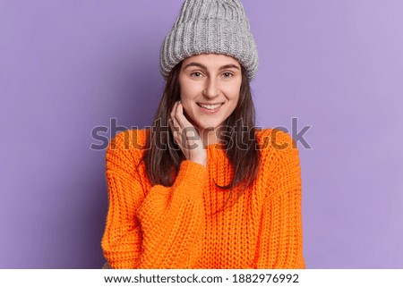 Photo of good looking millennial girl smiles pleasantly keeps hand near face dressed in knitted winter clothes has casual talk with friend glad to have weekend poses against purple background