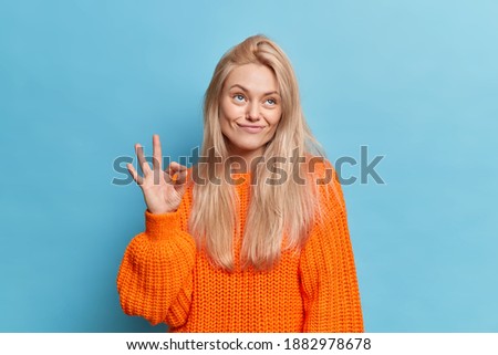 Photo of pleased toughtful young woman with natural blonde hair makes okay gesture stabds pensive wears knitted orange sweater isolated over blue background. People and body language concept