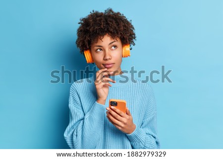 Photo of beautiful African American woman listens music with smartphone and headpones has thoughtful expression dressed in knitted jumpper isolated over blue background. Quarantine leisure concept Royalty-Free Stock Photo #1882979329