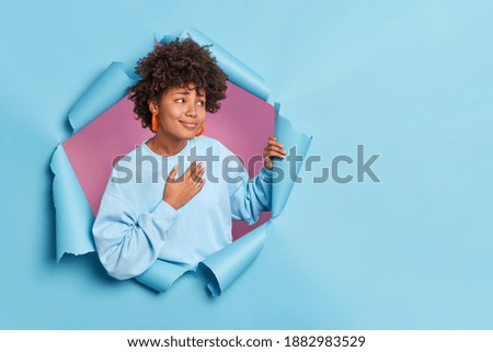 Pretty young African American woman presses hand to chest feels thankful expresses gratitude says thank you very much looks pleasantly aside dressed in casual jumper poses through hole of paper wall Royalty-Free Stock Photo #1882983529