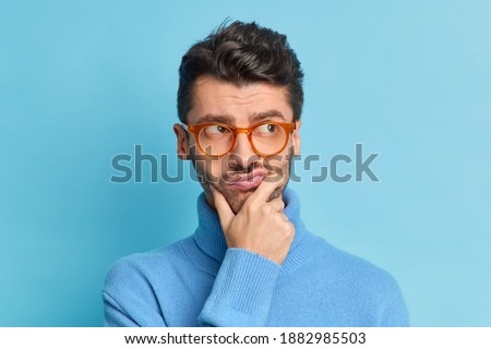 Headshot of displeased pensive adult man holds chin purses lips and looks aside tries to make decision wears spectacles and turtleneck isolated over blue background. People and thoughts concept Royalty-Free Stock Photo #1882985503
