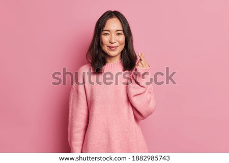 Pretty dark haired woman with gentle smile makes korean like sign expresses love to you wears casual long sleeved jumper isolated over pink background. Mini heart gesture. Body language concept