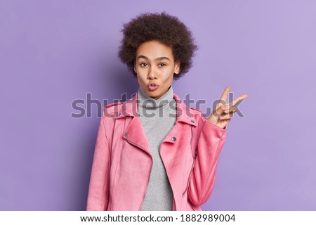 Pretty curly haired beautiful millennial girl makes peace gesture keeps lips folded shows v sign dressed in grey turtleneck and pink jacket isolated over purple background. Body language concept