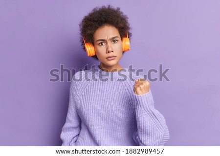 Irritated curly haired woman shows fist at camera expresses anger frowns face listens music uses stereo wireless headphones dressed in warm sweater isolated over purple background. Negative reaction