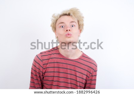 Shot of pleasant looking Young handsome Caucasian blond man standing against white background, pouts lips, looks at camera, Human facial expressions