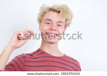 Young handsome Caucasian blond man standing against white background holding dental invisible aligner for tooth correction with a happy face standing and smiling with a confident smile showing teeth