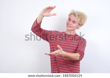 Young handsome Caucasian blond man standing against white background gesturing with hands showing big and large size sign, measure symbol. Smiling looking at the camera.
