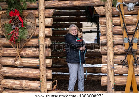 Older mature woman holds a yorkshire terrier dog while standing in a decorated log cabin hut