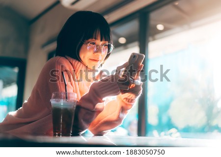 Young adult happy asian woman wear eyeglasses using mobile phone for social media application. Text online message at indoor cafe on day. Background with window and warm sunlight on winter season. Royalty-Free Stock Photo #1883050750