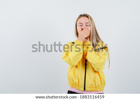  blonde girl holding hand on mouth in yellow jacket and looking sleepy. front view. 