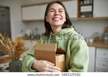 Cheerful teenage girl expressing positive emotions receiving unexpected birthday gift holding parcel and smiling with pleasure. Dark haired young woman posing with cardboard box with new cosmetics Royalty-Free Stock Photo #1883129182