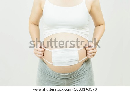 A tummy of a pregnant woman with medical face mask,Pregnancy health care preparing for baby concept.Motherhood among teenage mother, hugging belly, isolated on gray background