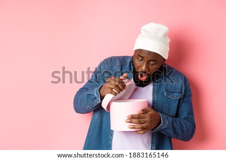Image of surprised african-american man open box with birthday gift, staring at present with amazement, standing against pink background