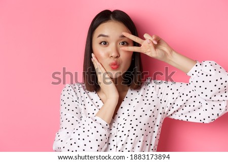Beauty and lifestyle concept. Close-up of pretty asian woman showing peace sign and touching cheek, smiling happy at camera, standing over pink background