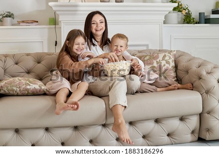 Mom sits on the couch with her son and daughter and watch a movie. A woman, a boy and a girl eat popcorn while watching a movie. The family is resting at home on the weekend