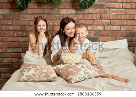 Mom sits on the bed with her son and daughter and watch a movie. A woman, a boy and a girl eat popcorn while watching a movie in the bedroom. The family is resting at home on the weekend